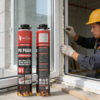 Two cans of WEIDU B1 foam in the foreground with a construction worker in the background wearing a yellow hard hat and gloves, applying insulating foam to a white window frame.